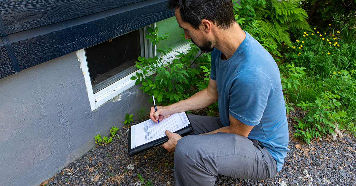 A home inspector writes something on his clipboard as he kneels down to inspect a basement window from the outside.