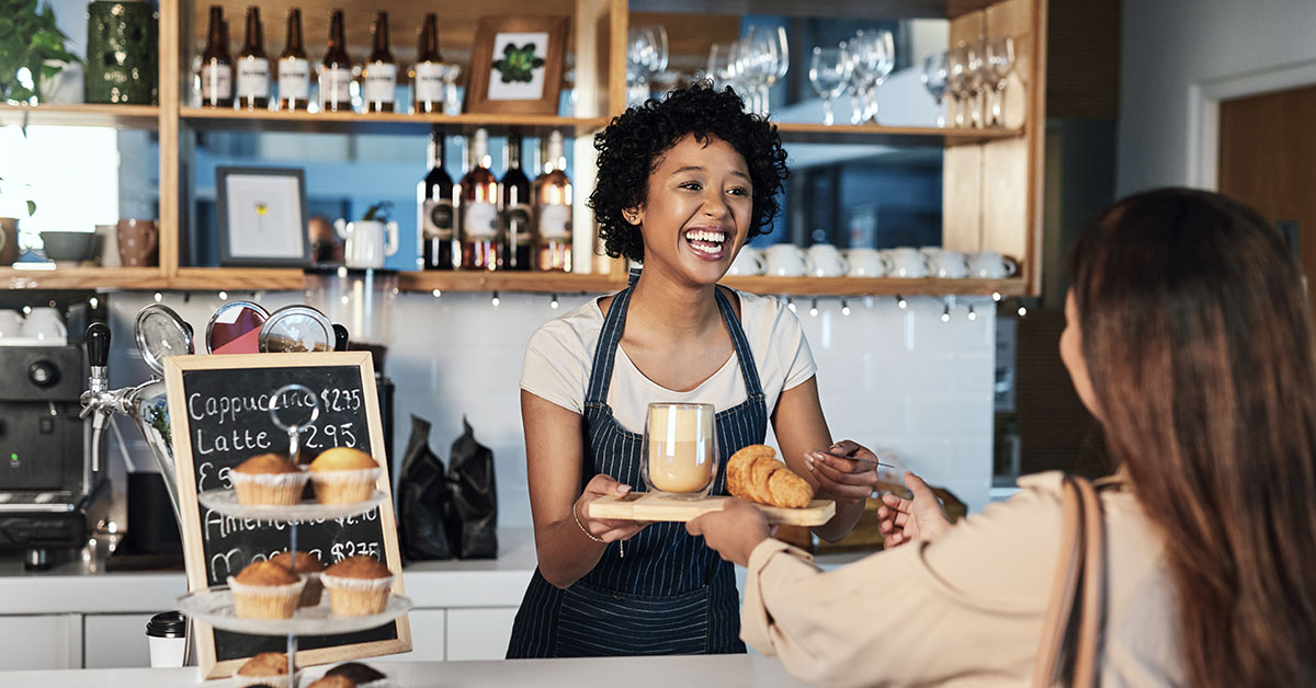 A female barista smiles as she passes a tray of food across the counter to a customer.