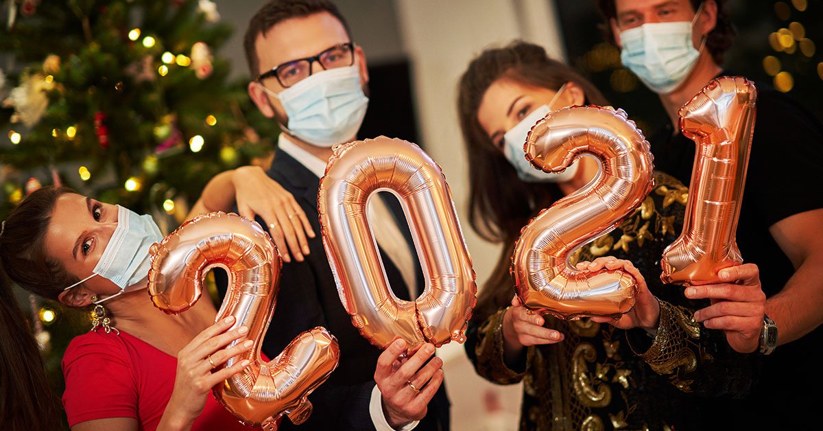 A group of young people wearing medical masks stands together and holds balloons that spells out 2021. There is a Christmas tree in the background.