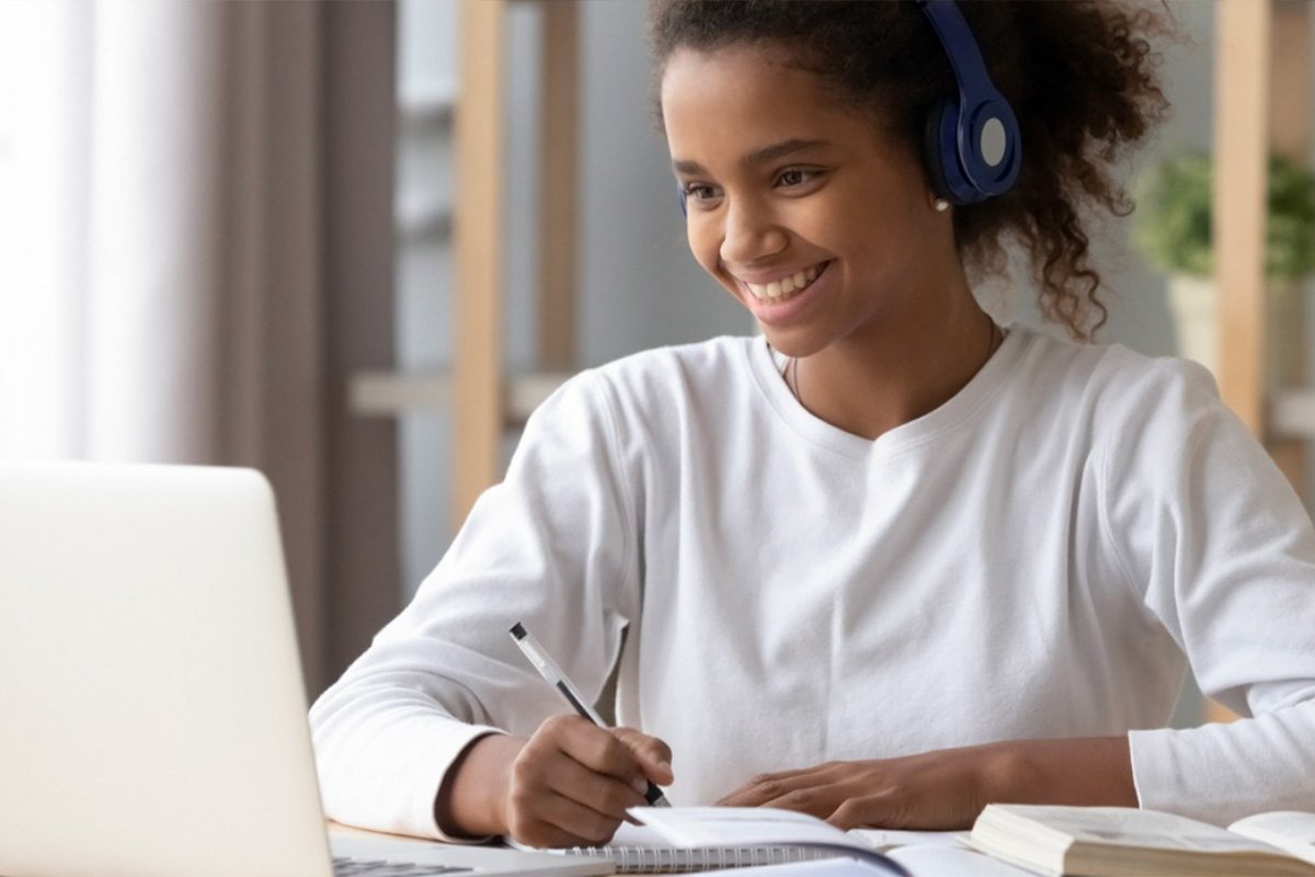 A smiling teen girl listening to music with headphones while looking at her laptop.