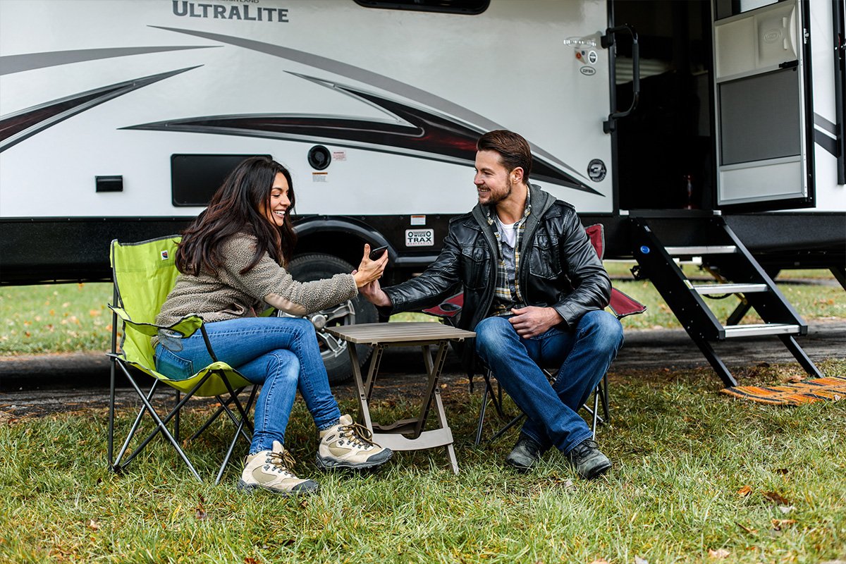 A smiling couple sitting next to their recreational vehicle looking at the phone.