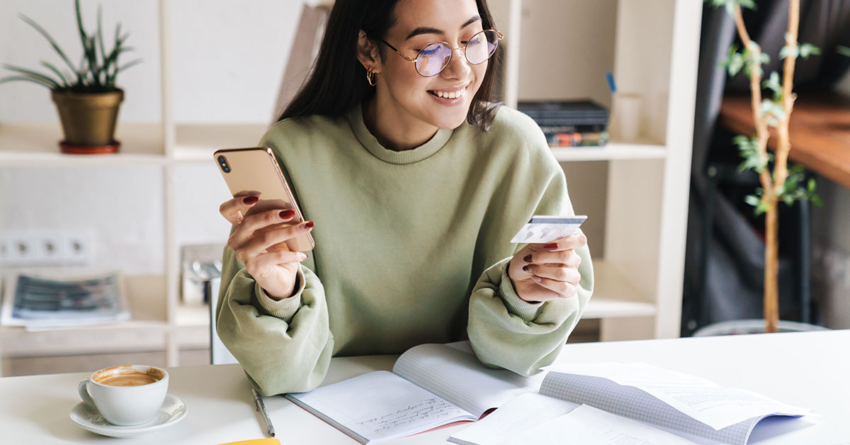 A young woman sits at a desk with papers and coffee. She is smiling and holding a phone in one hand and a credit card in the other.