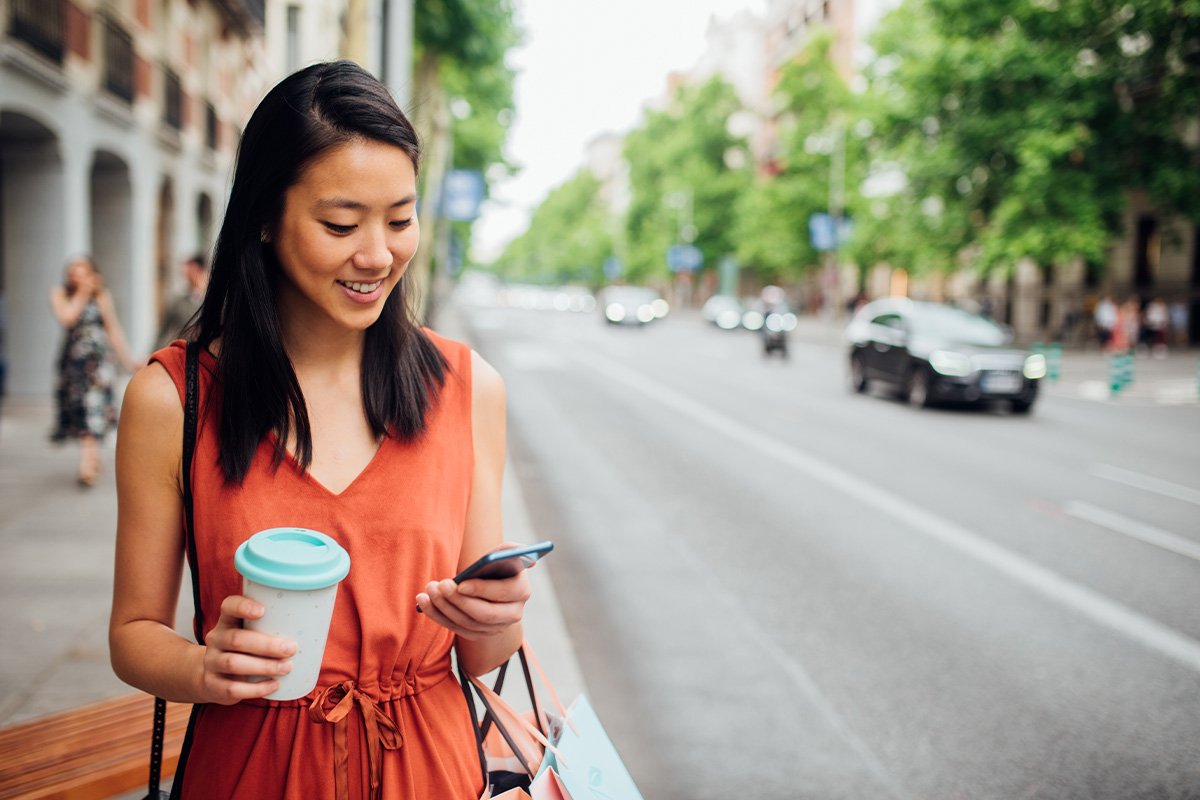 woman on the side walk smiling while holding a drink and a phone