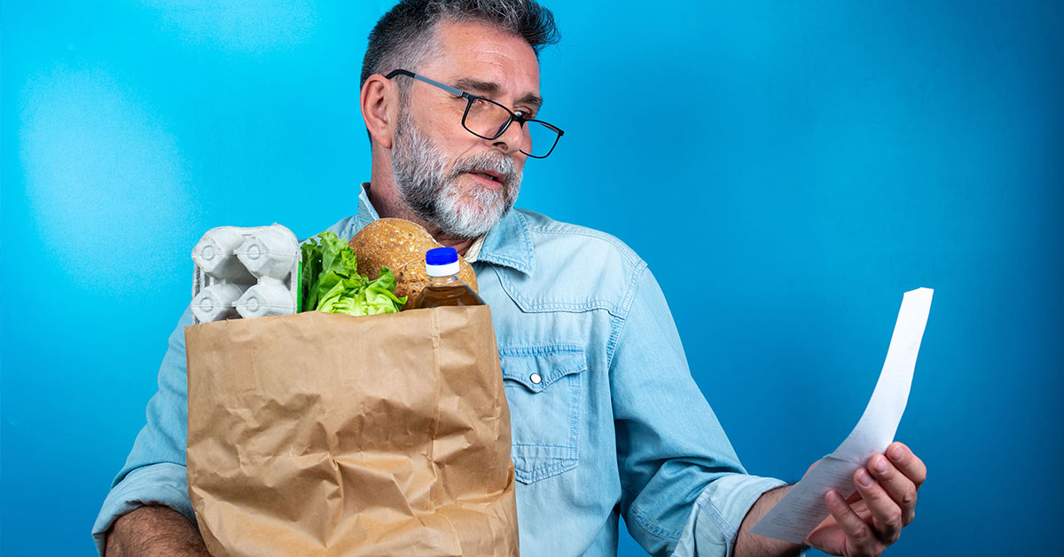 Older man looks shocked as he reads the receipt from his grocery run, while holding a paper bag of groceries in his right arm