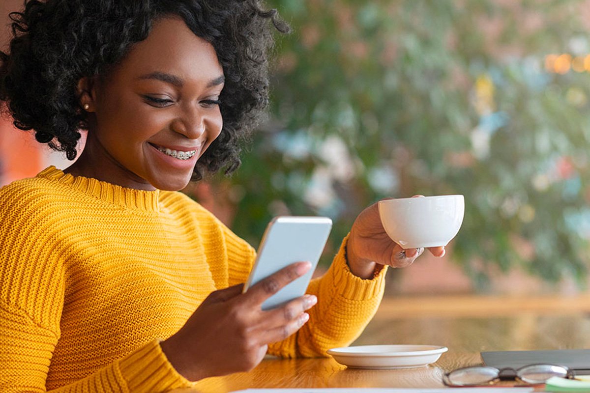 A girl smiling holding a cup and looking at her smart phone.