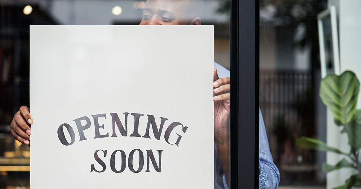 Person holding an “Opening Soon” sign up behind the glass door of his new business.