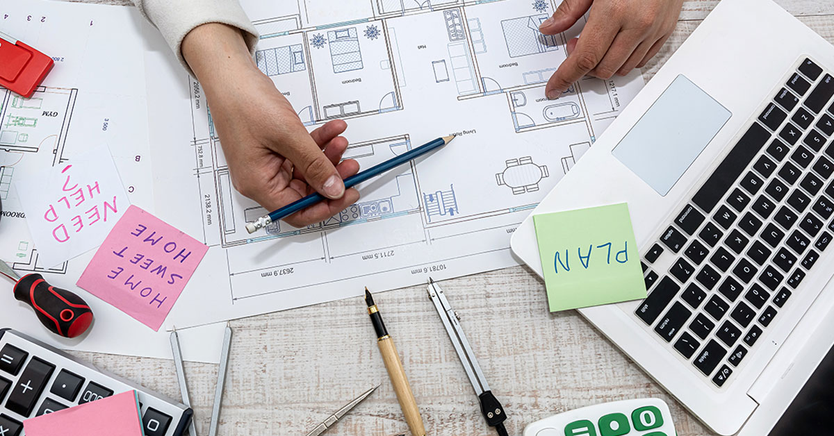 Photograph of homeowner reviewing home decoration plan on paper, with a laptop and two calculators on their desk.