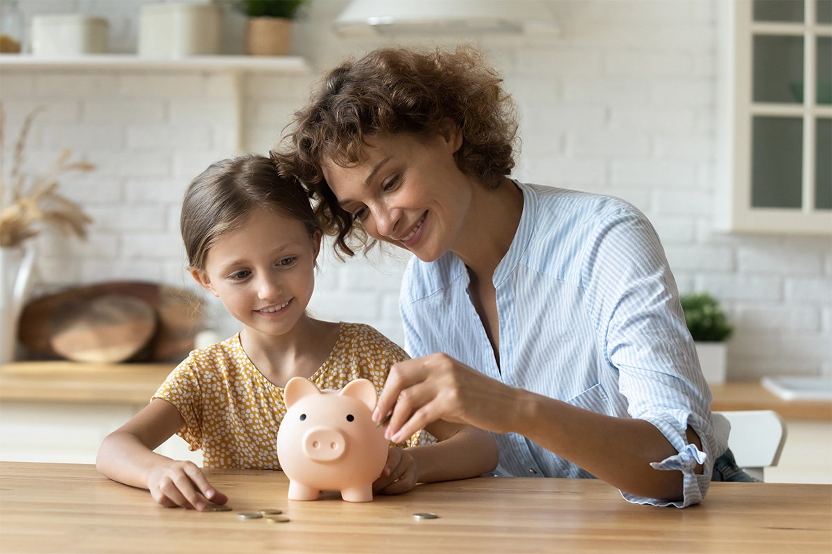 Mother with daughter in the kitchen putting coins into a piggy bank.