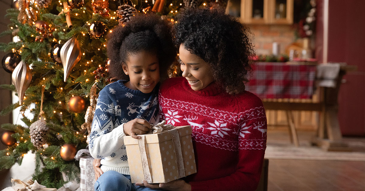 Daughter smiles as she sits on her mom’s lap and unwraps a Christmas present.